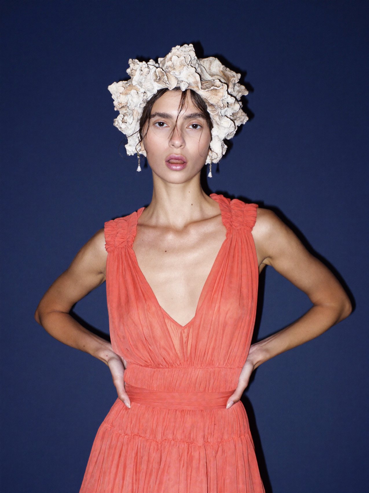 A model in a coral dress wearing a headpiece of oyster shells against a blue wall