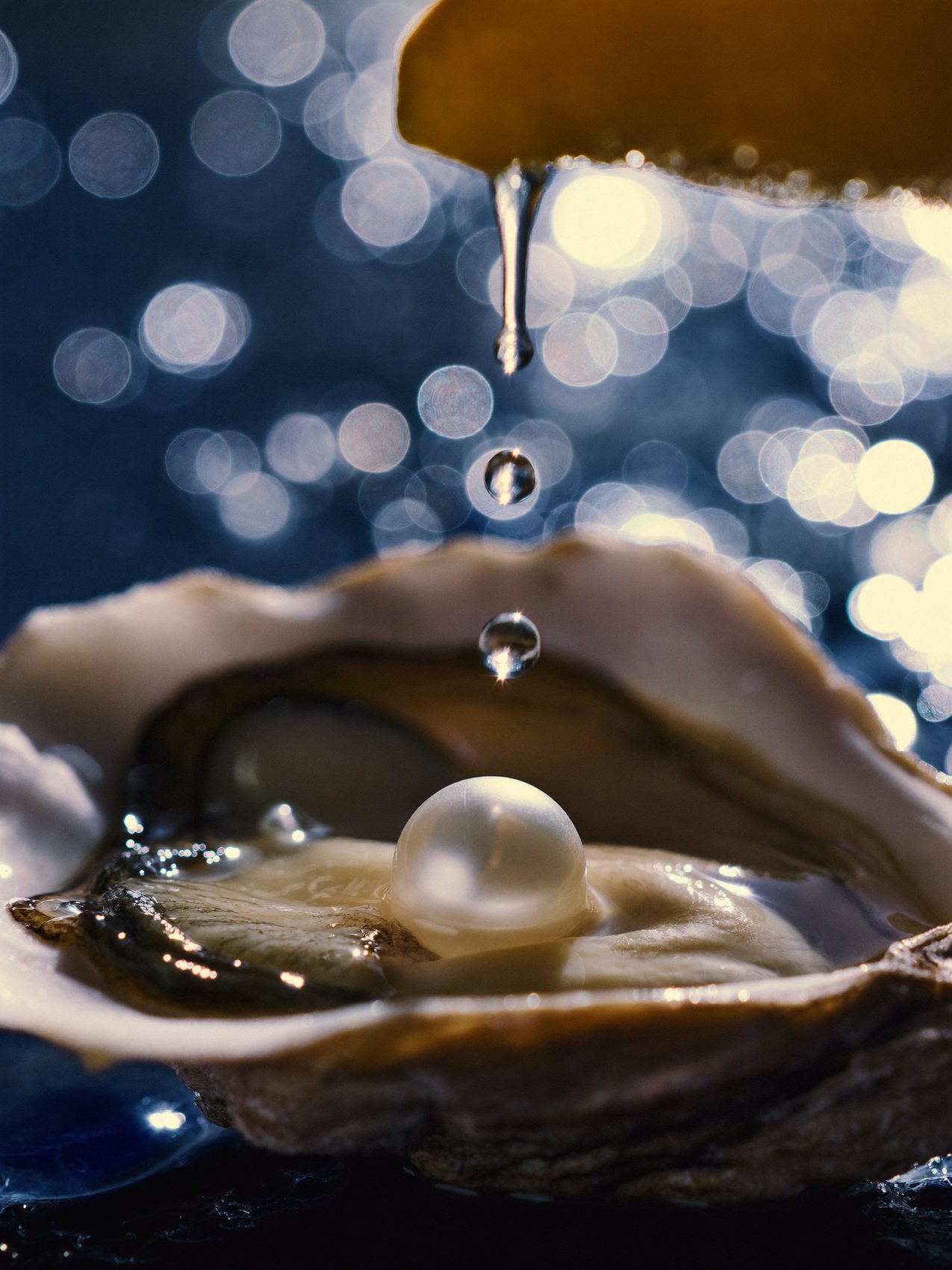 A pearl resting in an open oyster while a drop of lemon falls over it