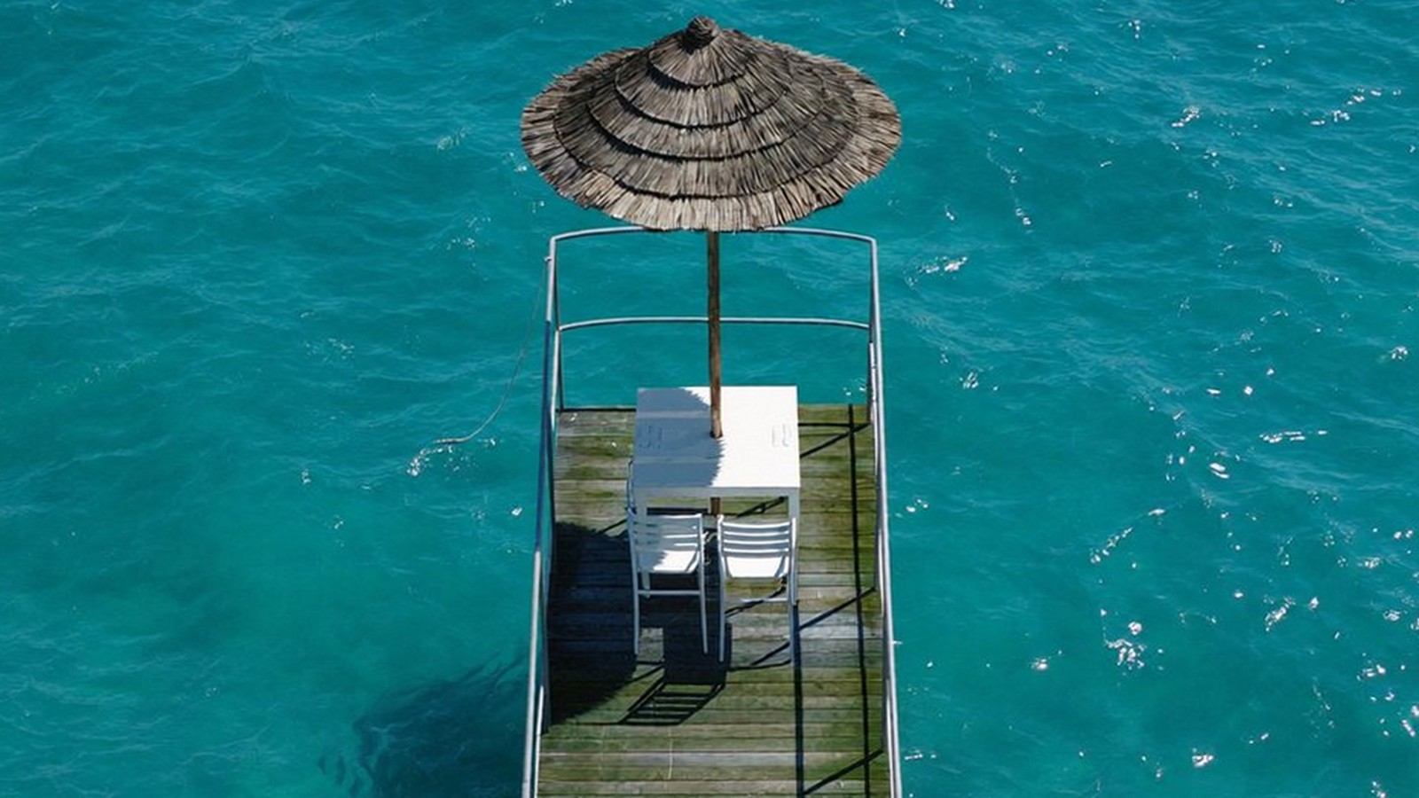 Aerial view of a wooden pier reaching into turquoise sea with a straw parasol at the end