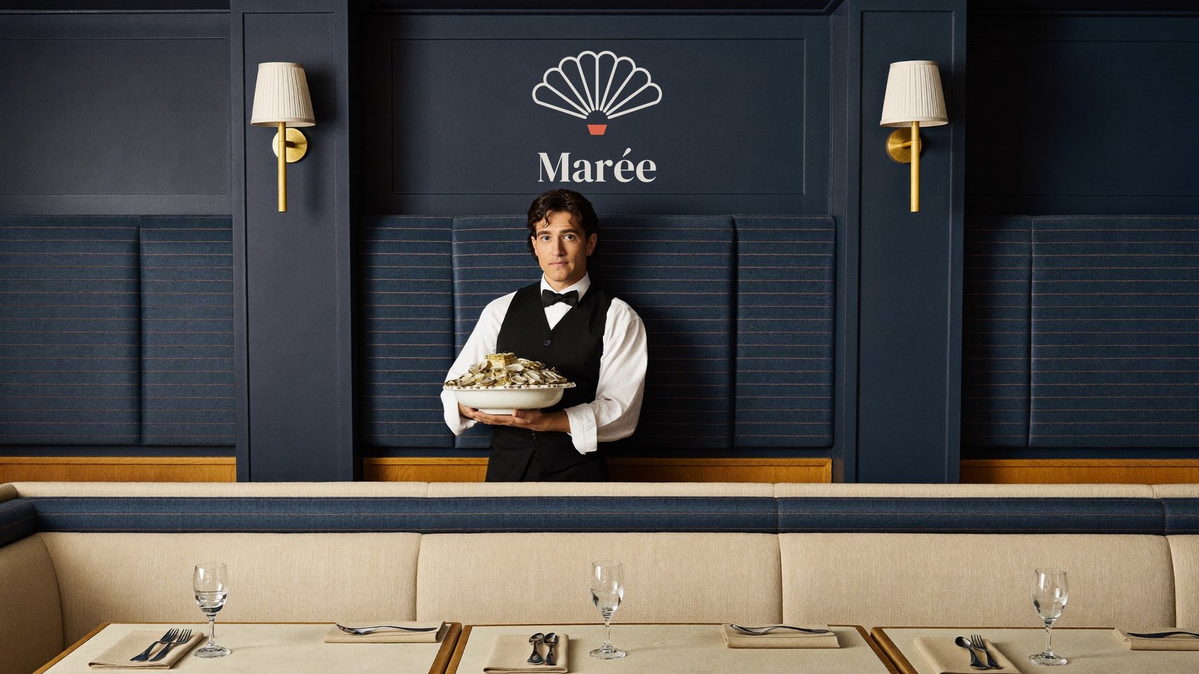 A waiter holds a platter of oysters in a navy dining room; the Marée mark is painted on the wall panel above him