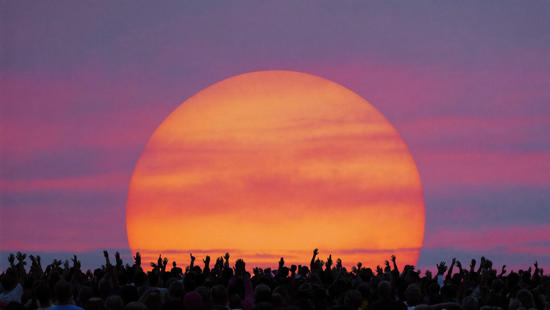 A crowd with raised hands in silhouette in front of an enormous orange sun on the horizon.