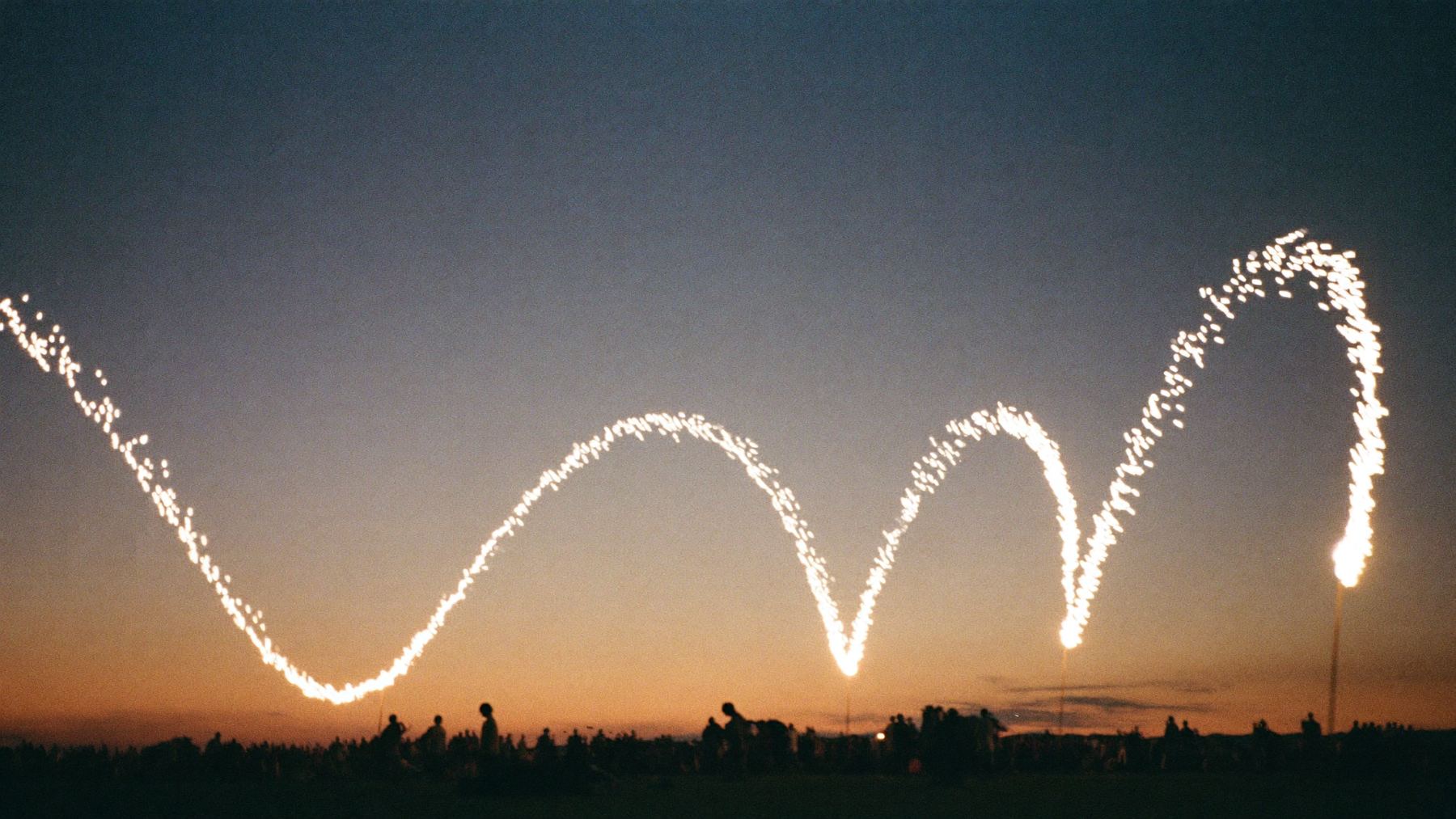 A line of sparks traces a looping path through a dusk sky above a field with a small crowd on the horizon.
