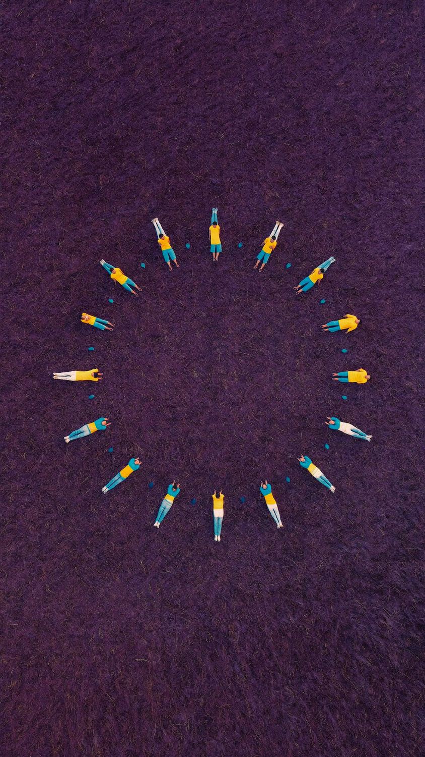 Seen from above, a dozen people in yellow and teal lie in a circle on dark purple ground, like the rays of a sun.