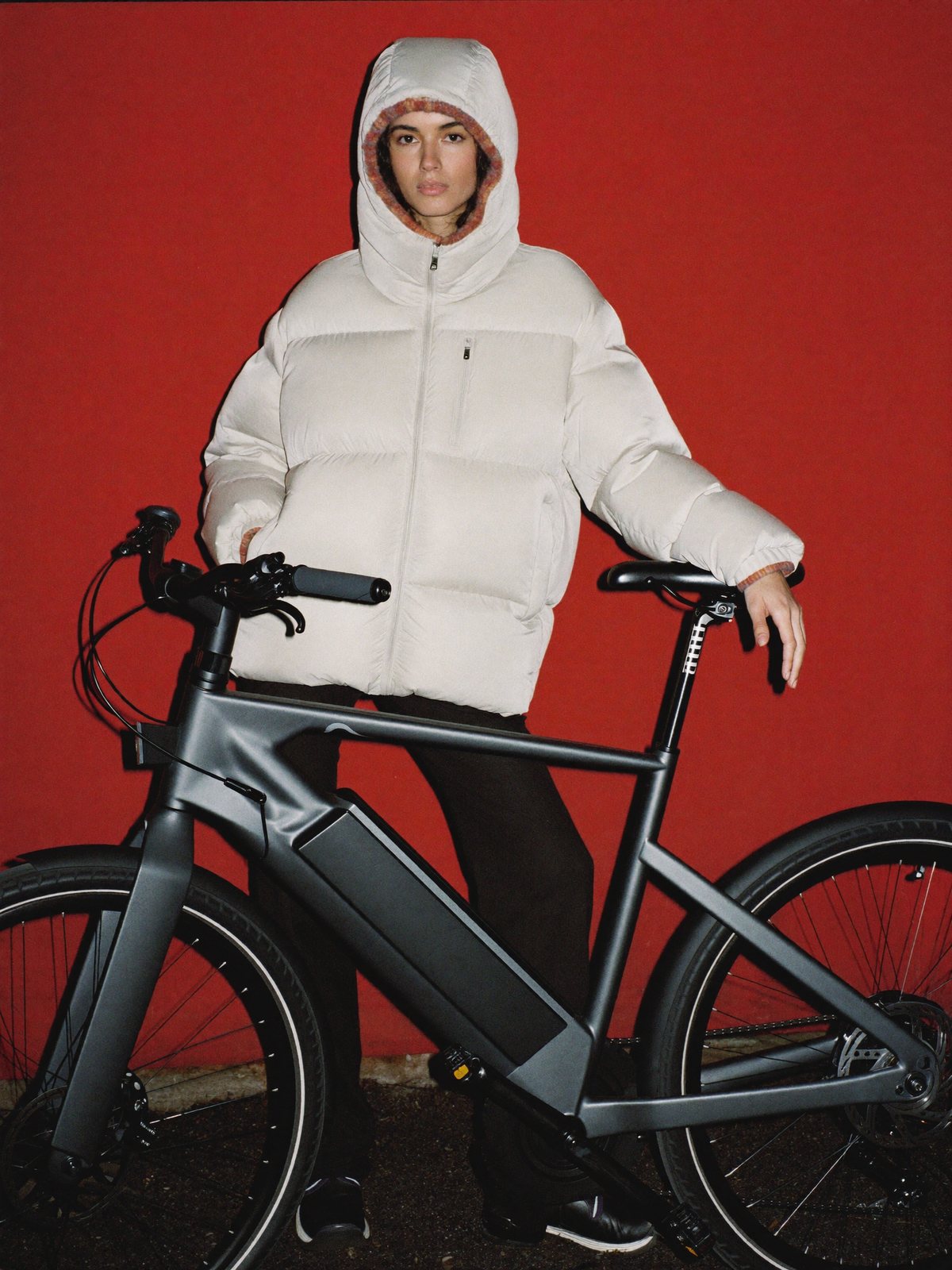 Woman in a white puffer jacket holding a grey e-bike against a red wall